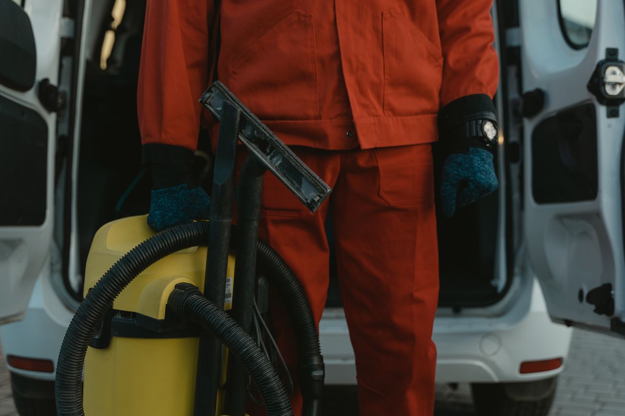 creative-03 A professional cleaner in a red uniform holding a vacuum near a van, ready for service.