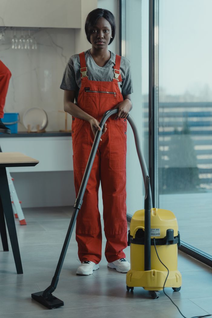 Services-02 An adult woman in red coveralls operates a vacuum cleaner in a modern indoor setting, focusing on cleaning tasks.